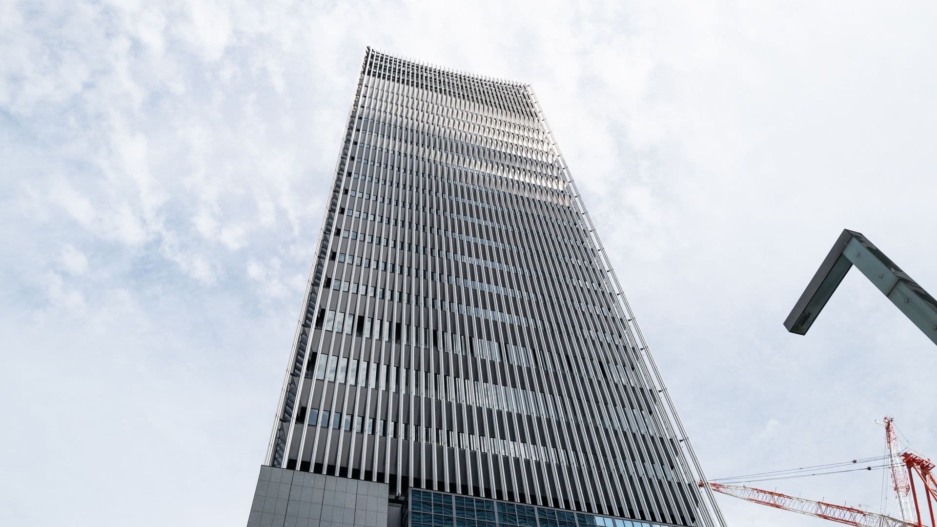 Low angle view of a tall modern skyscraper under construction with a crane in the background, against a cloudy sky.
