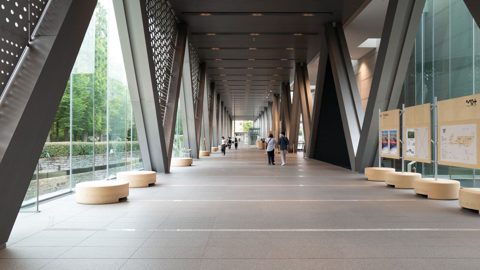 Modern indoor walkway with large glass windows, seating benches, and informational boards. A few people are walking and standing inside.