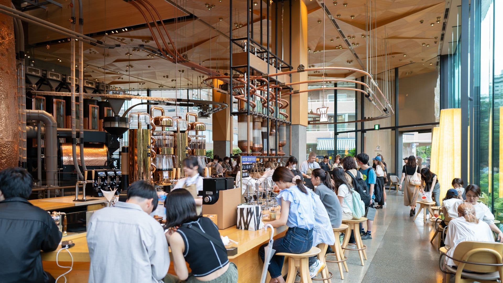 Inside a modern coffee shop with a busy counter, numerous customers sitting at bar stools and tables, large windows letting in natural light, and industrial-style decor with copper pipes and wooden accents.