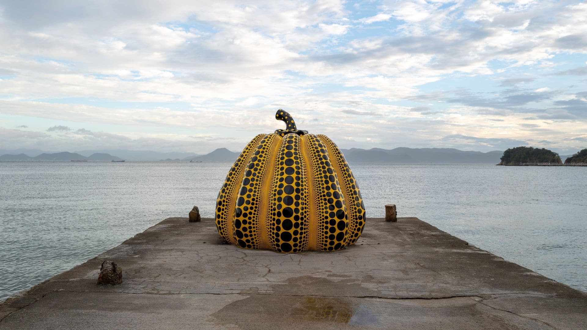 A large pumpkin sculpture painted yellow with black polka dots, placed on a concrete pier with water and distant islands in the background under a cloudy sky.