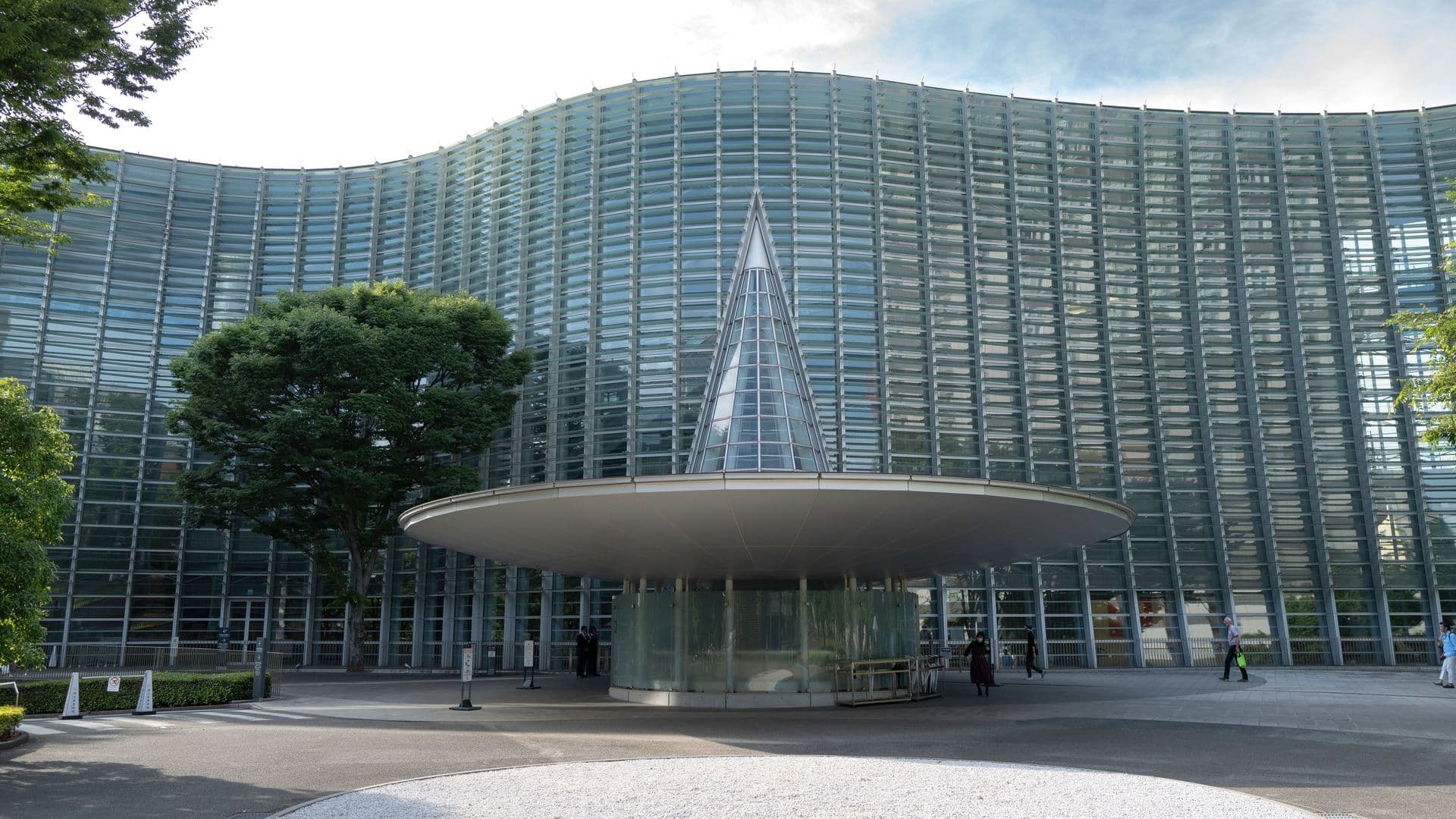 Modern glass building with curved facade, a triangular glass extension, and an outdoor structure resembling a large umbrella, with a few people walking nearby and trees on the sides.