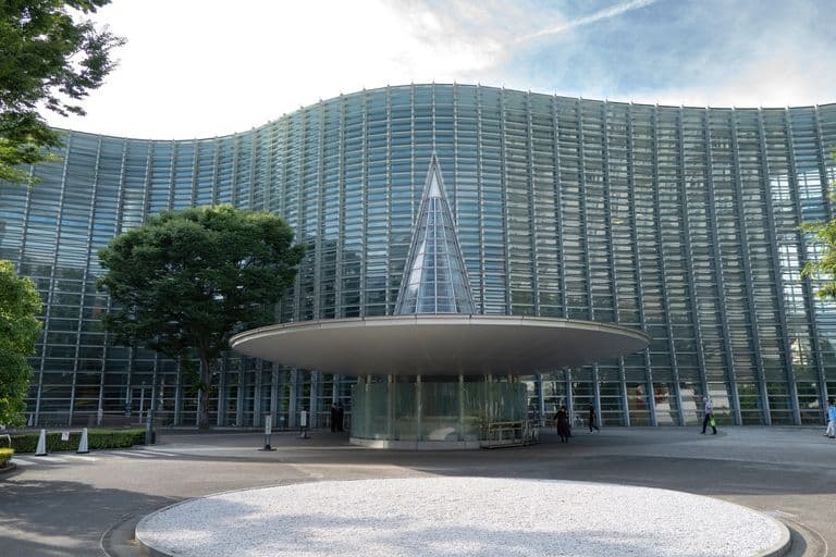 Modern glass building with a rounded entrance canopy and a glass cone structure, surrounded by a few trees and pedestrians.