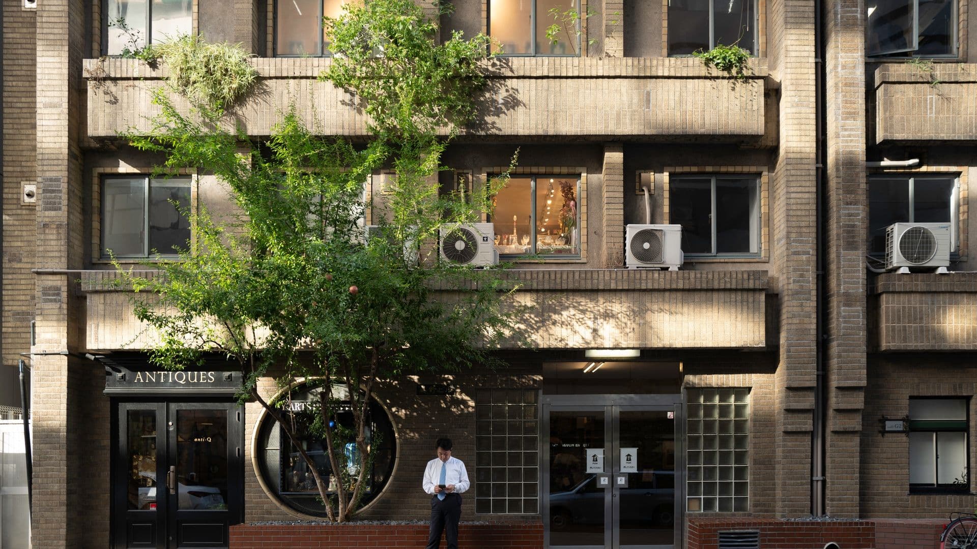 A man in business attire standing on the sidewalk outside a brick building, looking at his phone. The building has multiple windows, some with air conditioning units, and a storefront with an antique shop sign. A large leafy tree partially obscures the building's facade.