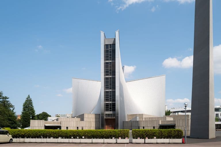 Modern church building with white metallic exterior, tall central tower, and curved wings flanking the tower, set against blue sky with some clouds and surrounded by greenery.