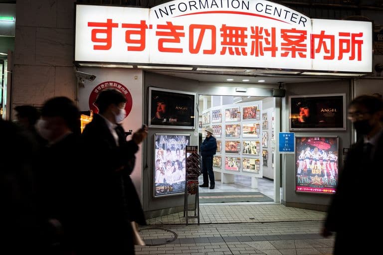 People walking past an illuminated entrance to an information center with Japanese signs, advertisements for entertainment, and a person inside browsing displayed posters.