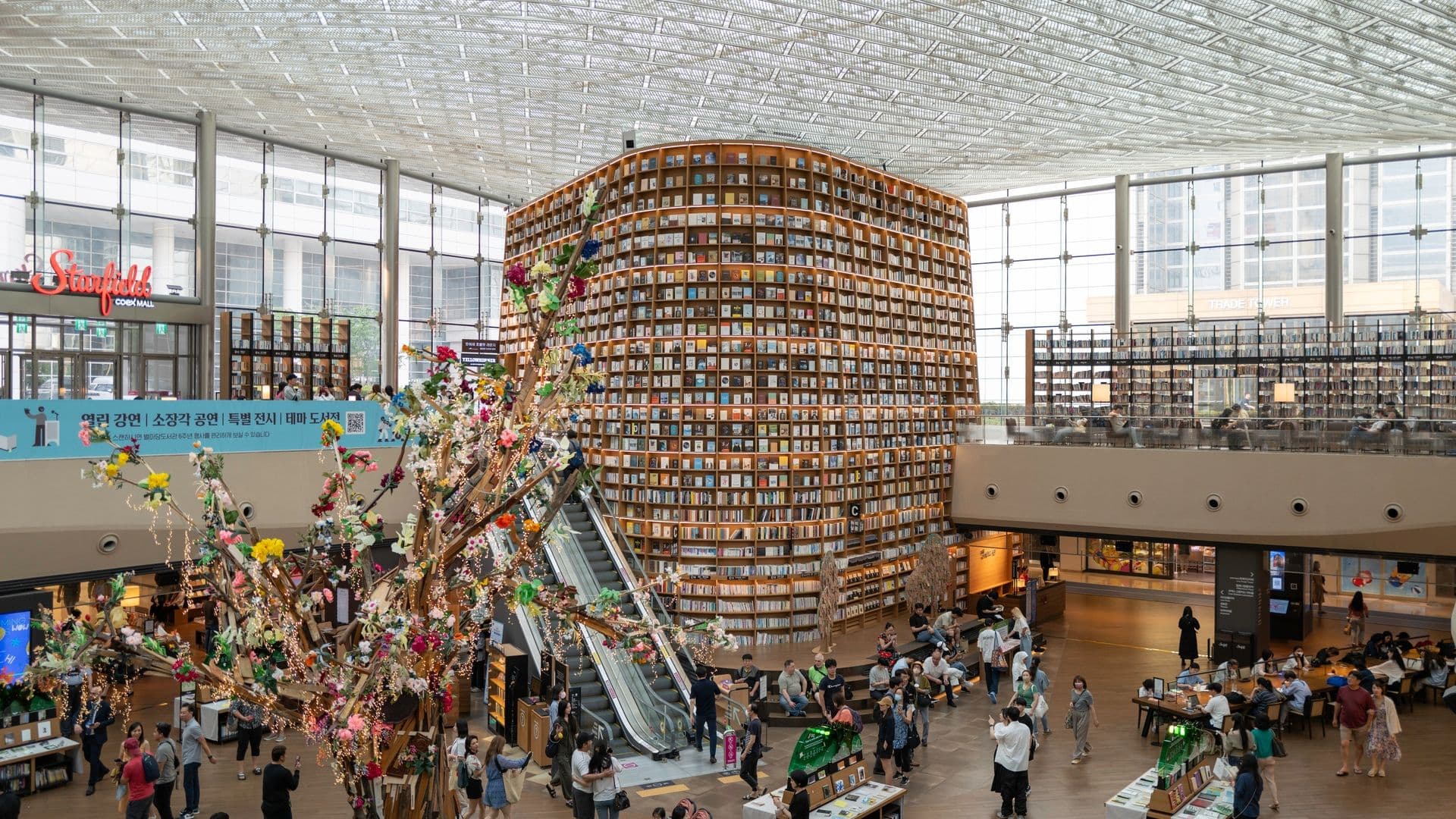 Interior view of a large modern bookstore with tall round bookshelves, a decorative tree in the center, escalators, and multiple visitors browsing and sitting around.