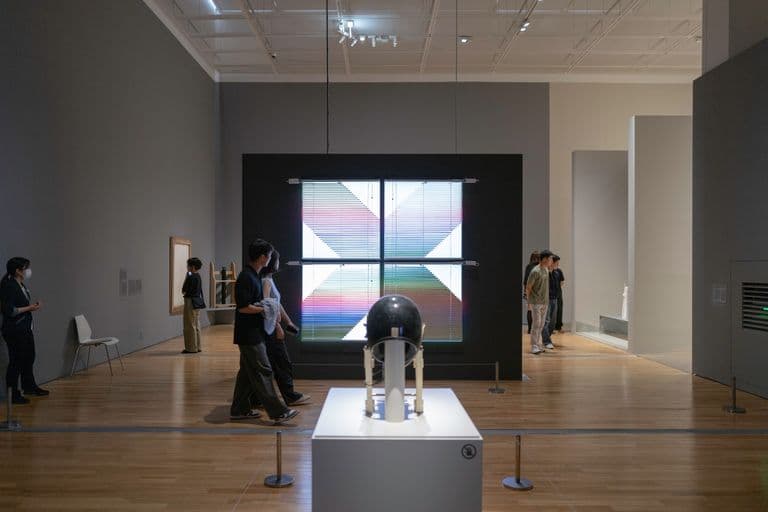 People viewing art and science exhibits in a museum, including a large colorful geometric artwork on the wall, a black spherical object on a white pedestal, and small sculptures on display.