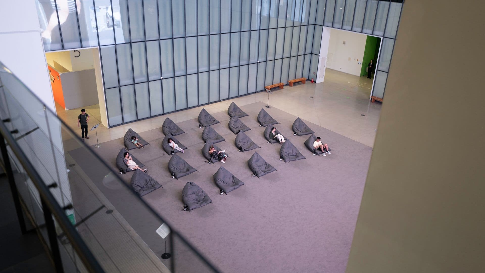 People sitting on bean bag chairs in a modern, spacious indoor lounge area with glass walls and benches, spaced apart and wearing face masks.