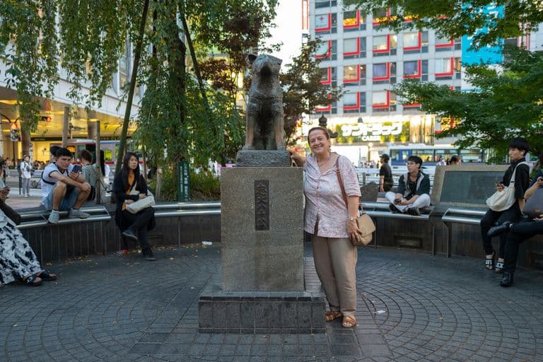 A woman standing next to a dog statue in a city park with people sitting on benches and modern buildings in the background.