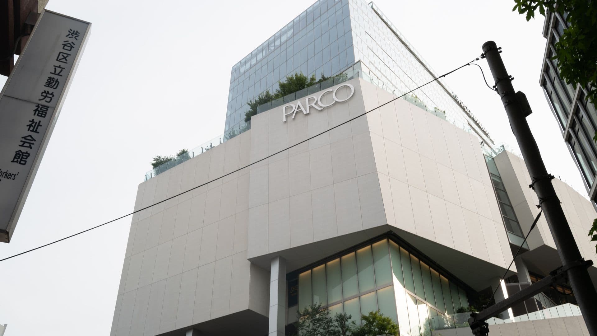 Modern multi-story building with the sign "PARCO" on it, featuring large glass windows and outdoor greenery at street level, viewed from a low angle.