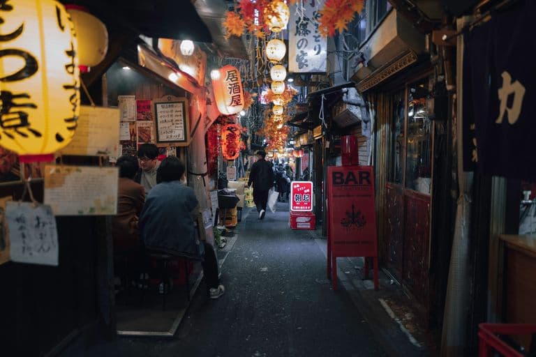 A narrow Japanese alleyway lined with small restaurants and bars decorated with lanterns and colorful signs, with people sitting outside and walking through.