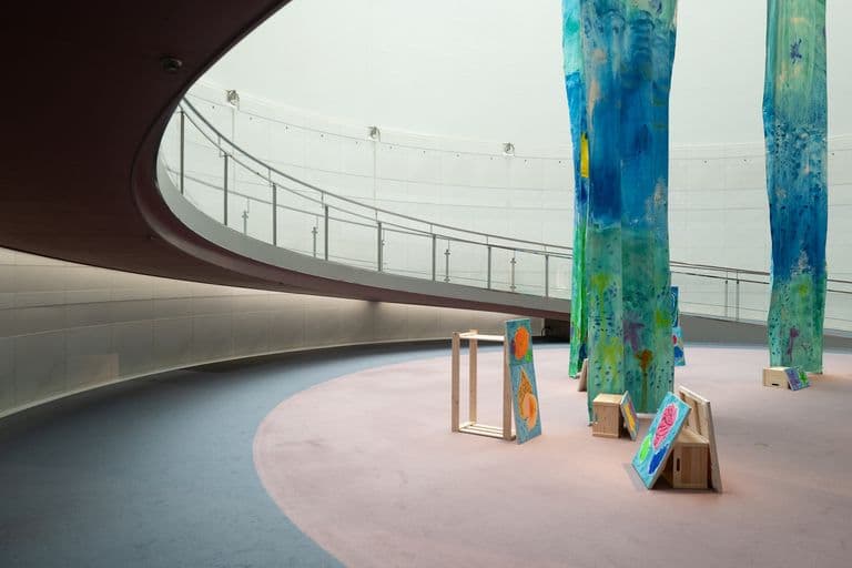 Art installation inside a modern building featuring tall, abstract, painted fabric sculptures and small painted canvas artworks on wooden easels and boxes.