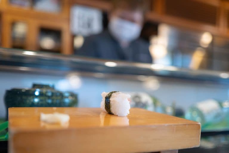 A piece of sushi with white rice and seaweed on a wooden sushi board, with a blurred sushi chef in the background.