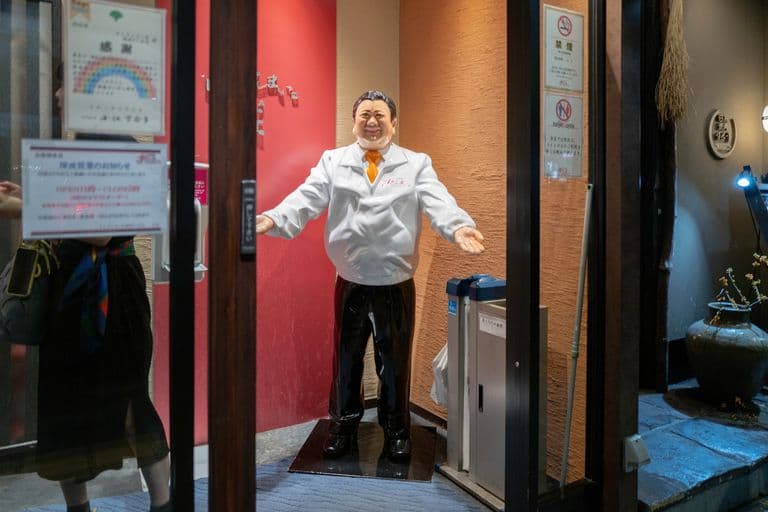A life-sized statue of a man dressed as a chef with a white coat and dark pants, standing with open arms in the entrance of a restaurant.