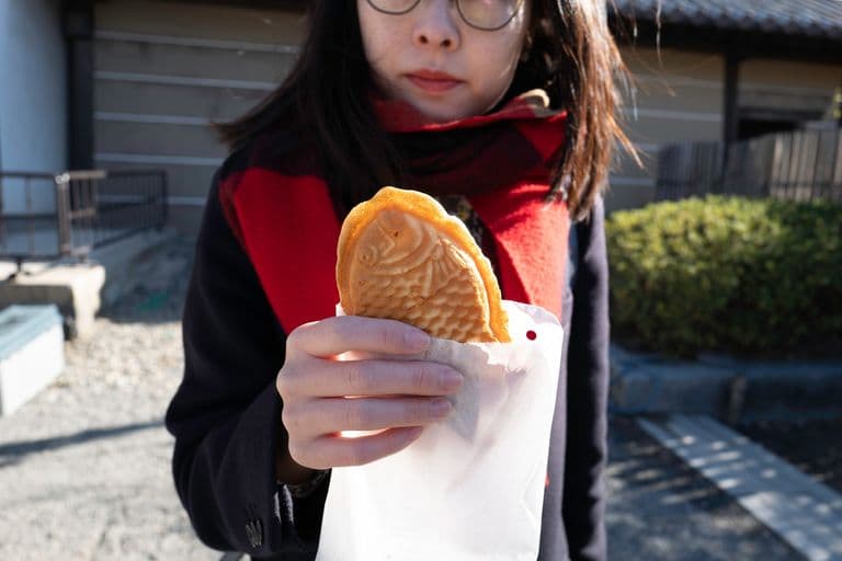 A woman holding a taiyaki fish-shaped pastry in a paper bag outdoors.