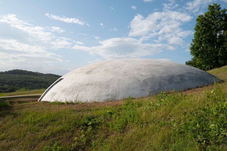 A large, white, dome-shaped structure on a grassy hill with trees and a blue sky with scattered clouds in the background.