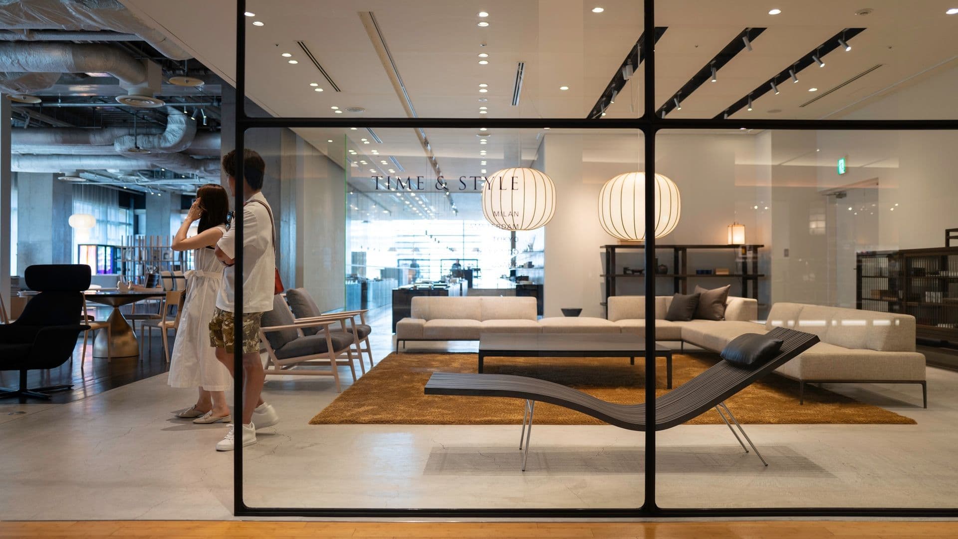 Modern furniture showroom with beige sofas, a black lounge chair, and large hanging paper lanterns. Two women are standing and talking inside, with the store name 'Time & Style' visible in the background.