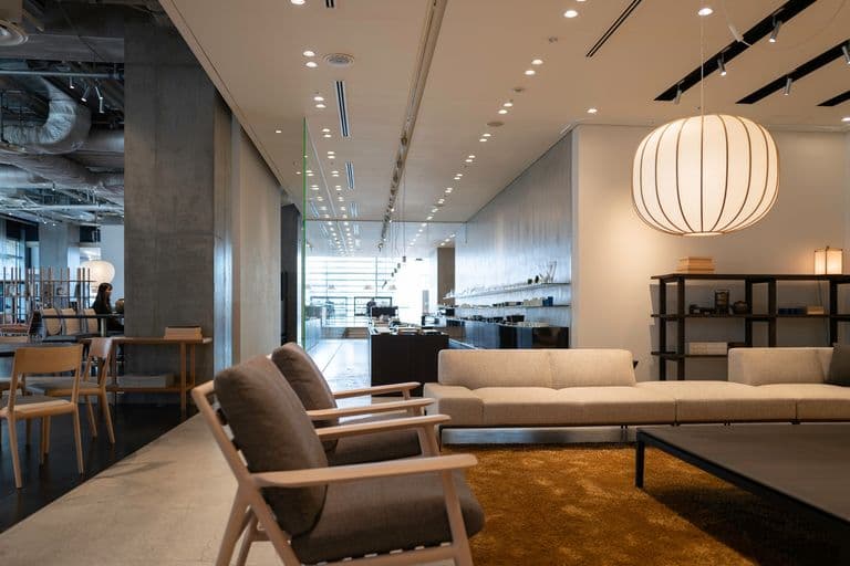 Modern hotel lobby with beige sectional sofa, brown armchairs, black coffee table, large white paper lantern pendant light, wooden shelving, and a carpeted area, with a bright outdoor area visible through large glass doors.