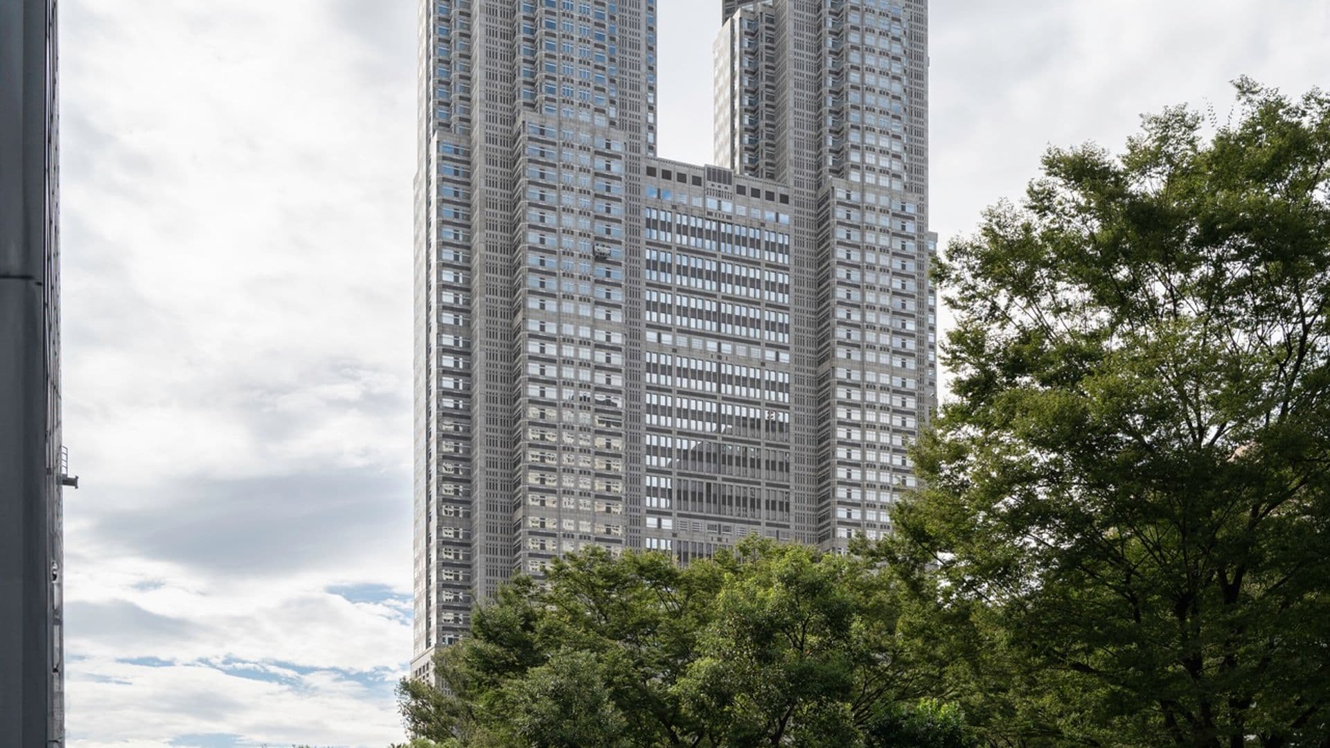 A tall modern skyscraper with two towers connected at the base and at the top, featuring geometric designs with round accents, surrounded by trees and a few people walking or sitting in a city plaza with stairs.