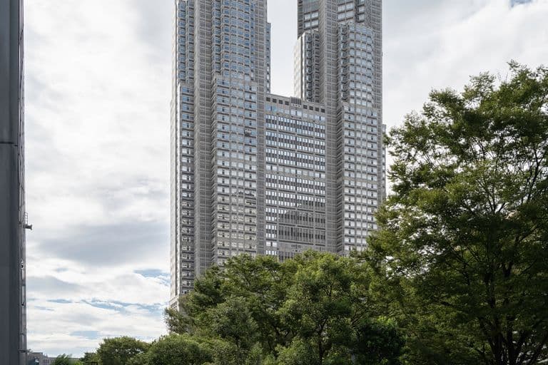 Tall modern skyscraper with a unique geometric design in an urban area, surrounded by trees and a tiled walkway with people walking.