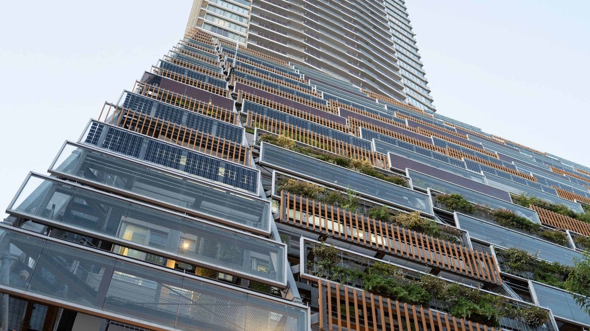 Tall modern skyscraper with glass balconies and greenery on the lower floors, viewed from below against a clear sky.