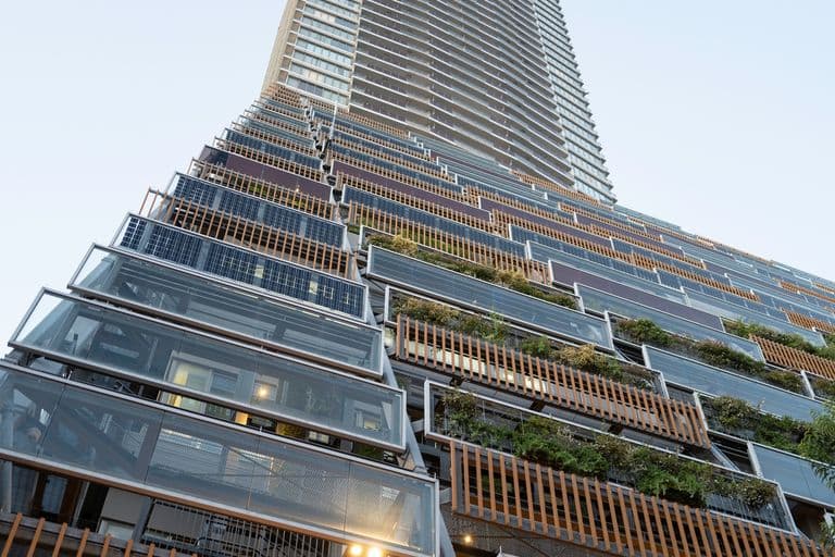 Low-angle view of a modern skyscraper with glass balconies and greenery on some levels against a clear sky.