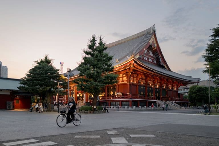 A traditional Japanese temple lit up at dusk, surrounded by trees and people, with a cyclist riding in the foreground and a cityscape in the background.