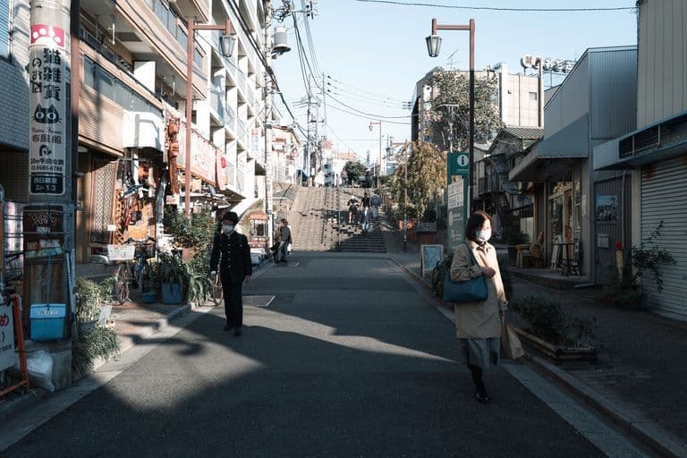 Street scene with pedestrians wearing masks, narrow sidewalk, small shops, stairs leading up the hill, utility poles and wires, clear weather, daytime.