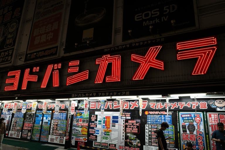 Brightly lit storefront with neon Japanese signage and advertisements, with a large red neon sign and multiple posters displaying electronics and promotions, at night.