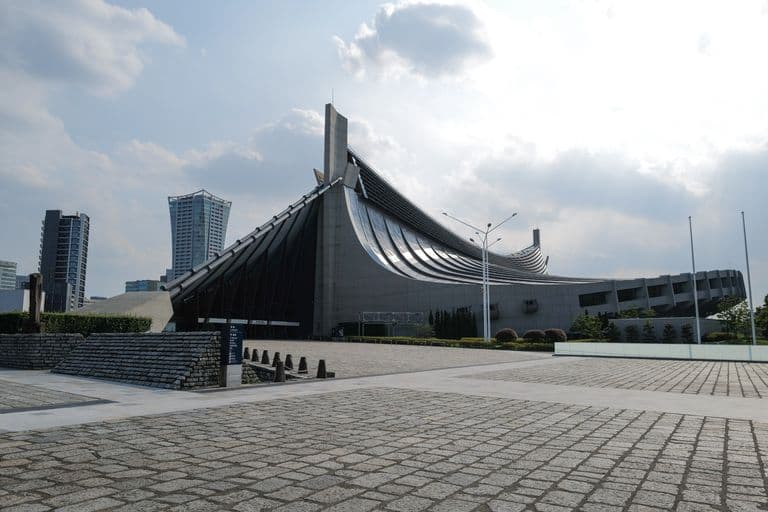 Modern building with an unusual, curved architectural design, surrounded by paved open space, with some high-rise buildings in the background under a partly cloudy sky.