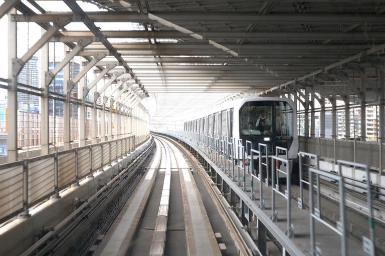 Modern train traveling on elevated tracks inside a partially enclosed station with city buildings in the background.