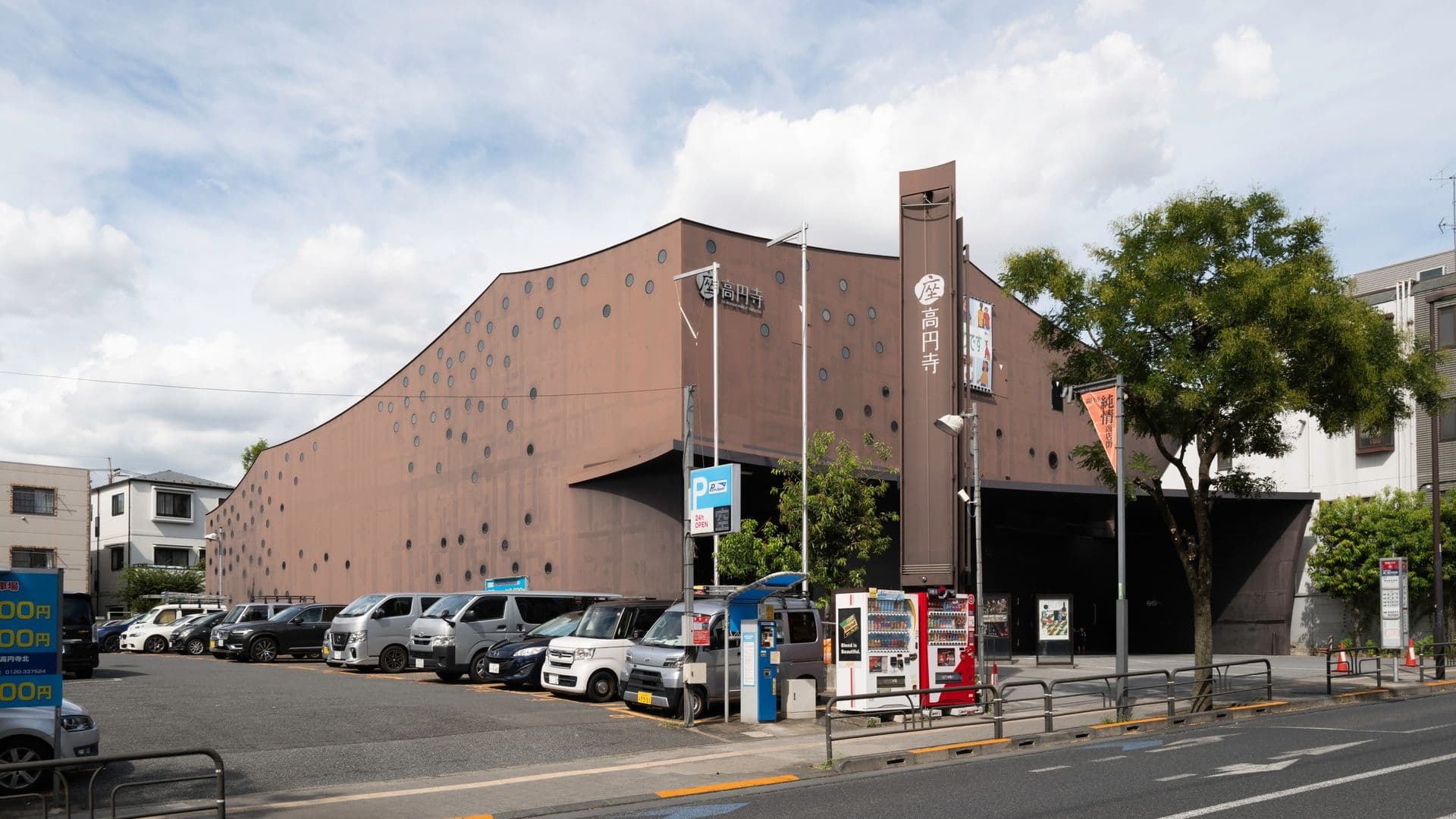 A modern brown building with circular patterns and Japanese signage, parking lot with cars, vending machines, trees, and a street with signs and railings.