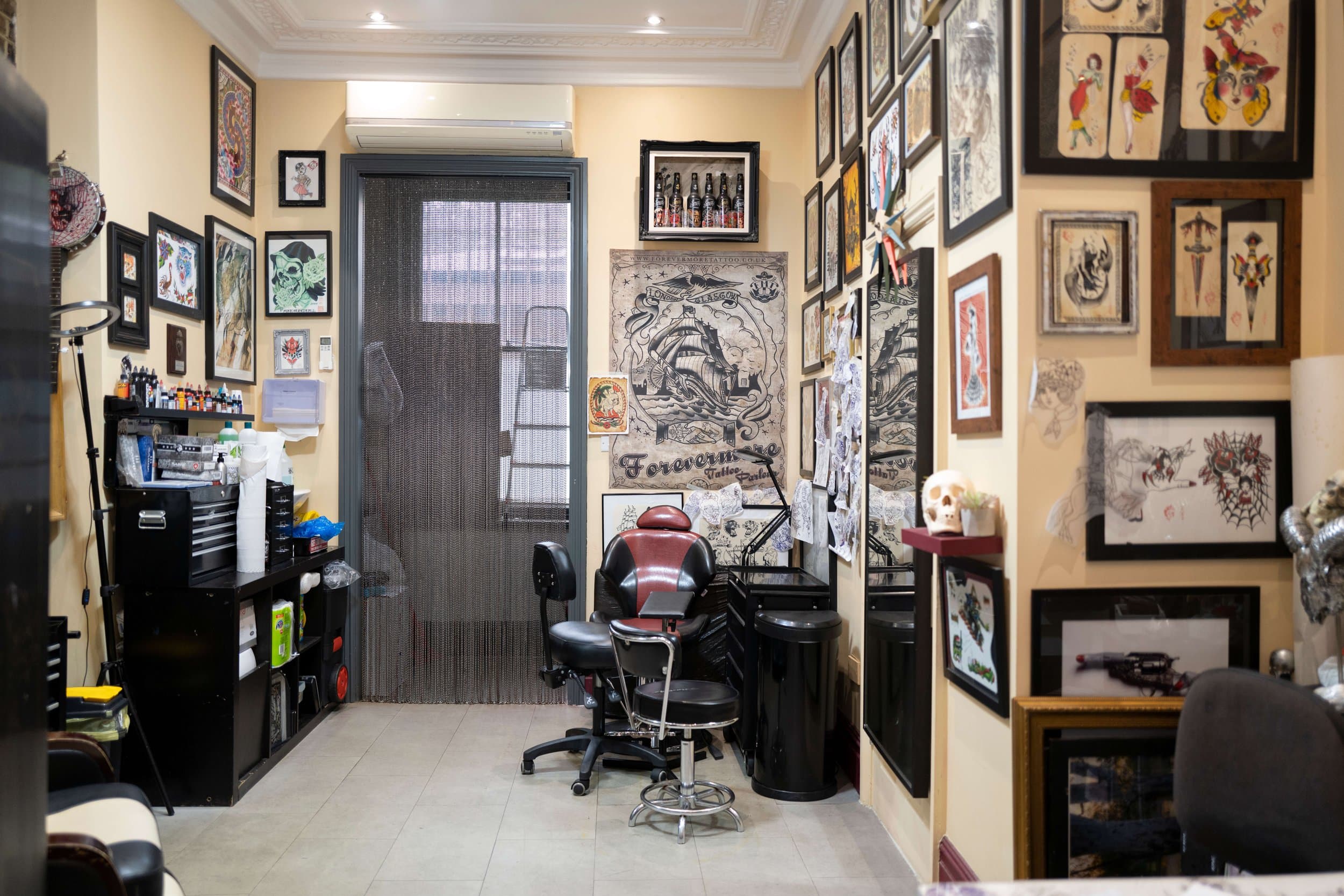 Tattoo studio with framed artwork on the walls, tattoo supplies on shelves, chairs, and a large tattoo-themed poster.