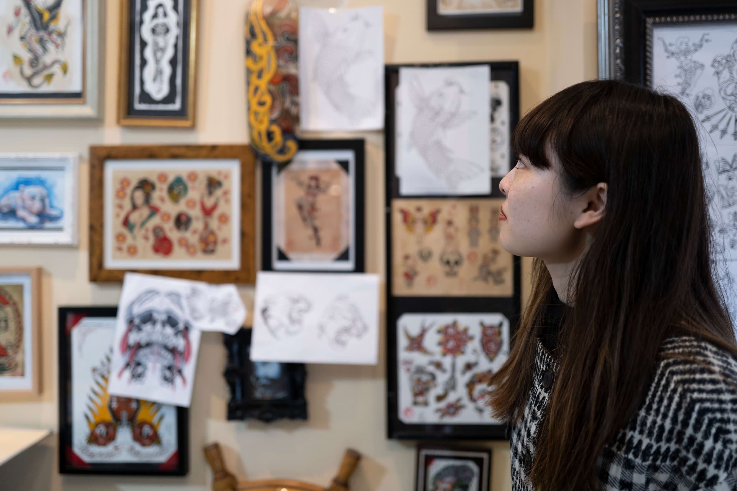 A woman with long dark hair looking at a wall filled with framed tattoo flash art and sketches in a tattoo studio or art gallery.