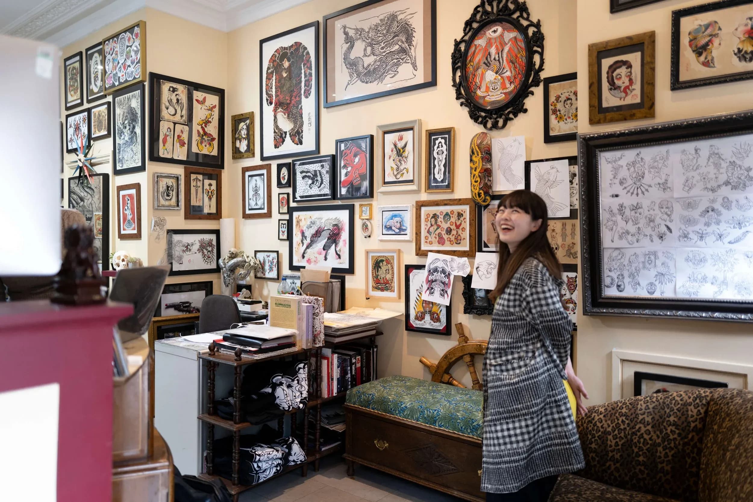 A young woman standing in an art studio or gallery with a smile, surrounded by numerous tattoo-style artwork and framed pictures on the walls.