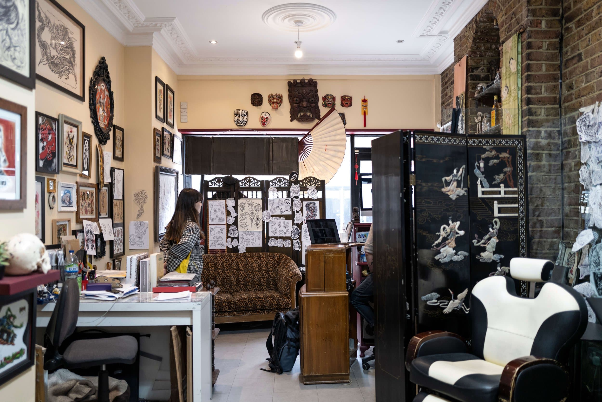 Interior of an art studio or gallery with various Asian-themed artworks and masks on the wall, a woman inspecting art, and folding screens with painted figures.