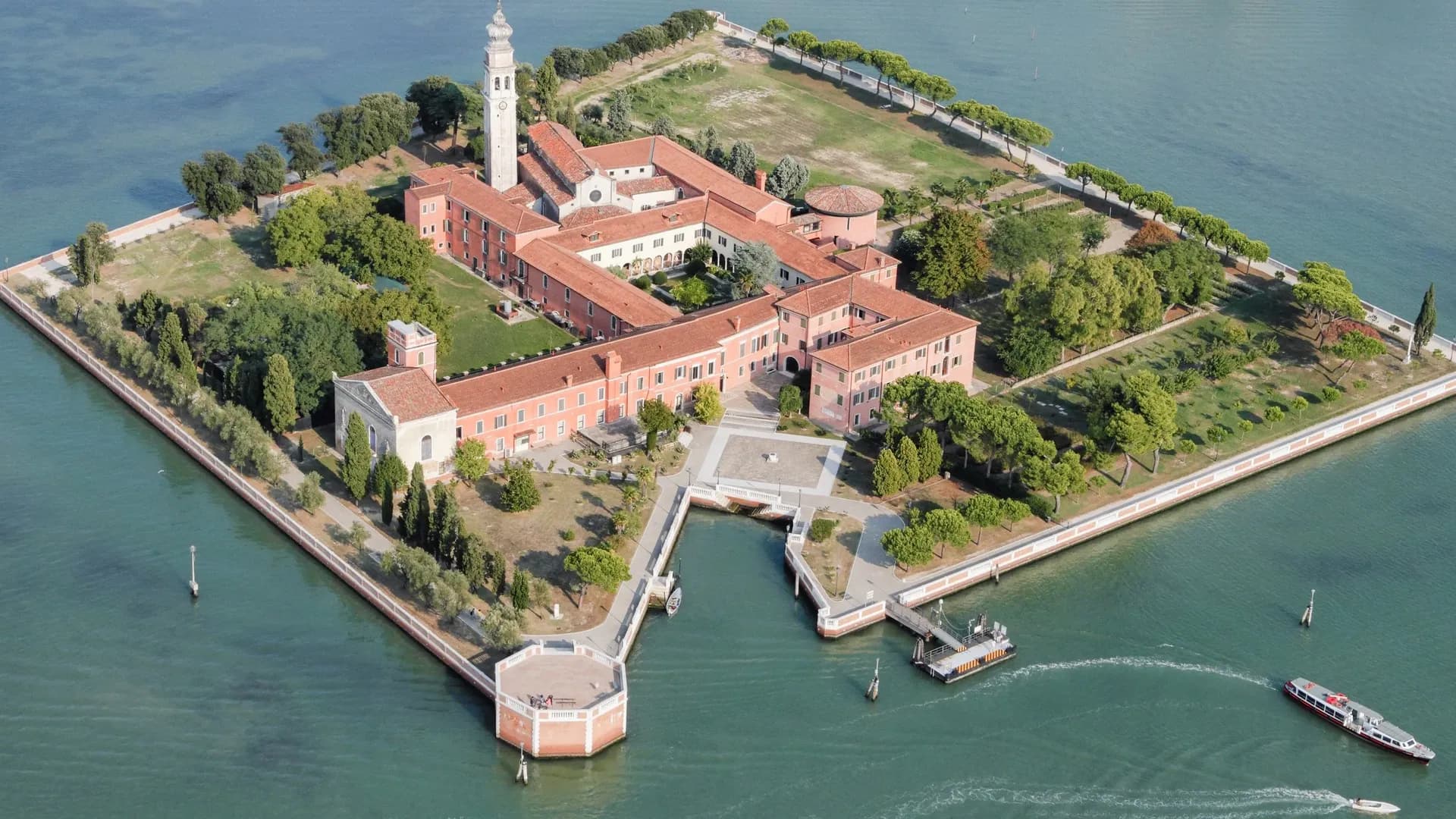 Aerial view of a square-shaped island in Venice with historical buildings, a tall clock tower, courtyards, and surrounding gardens. Boats are traveling in the surrounding water.