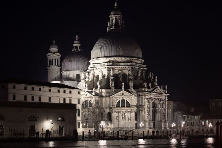 Night view of a grand historic cathedral with domes and sculptures, located by a waterway with reflection, lit by streetlights.