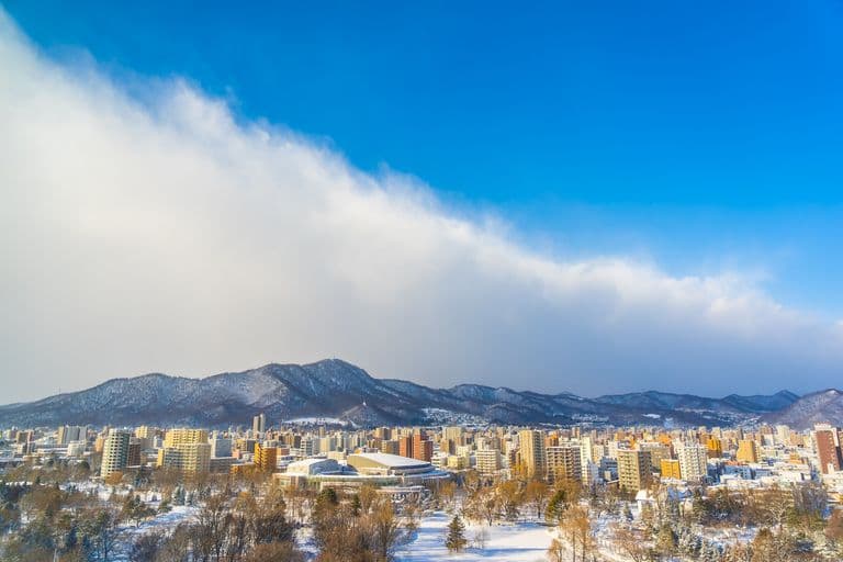 Snow-covered cityscape with mountains in the background and a partly cloudy blue sky.