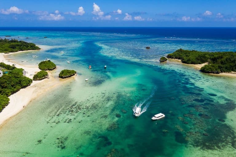 Aerial view of a tropical beach with boats on turquoise waters, sandy shores, green islands, and a blue sky with clouds.