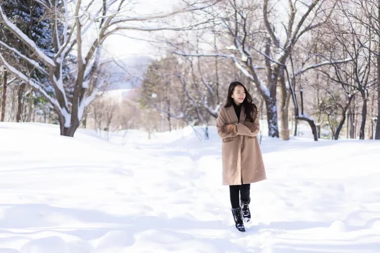 A woman in a tan coat and black boots walking through a snowy park with trees covered in snow, smiling and looking to the side.