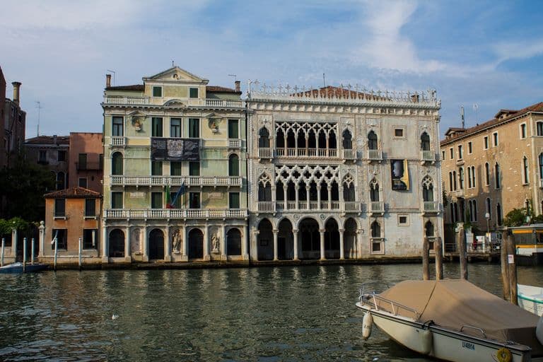 Two ornate historic buildings along a canal in Venice, Italy, with a boat in the foreground and a cloudy sky above.