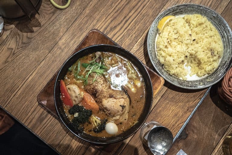 A bowl of ramen soup with carrots, broccoli, lotus root, chicken, and garnished with green onions on a wooden tray, served with a bowl of rice on a wooden table.