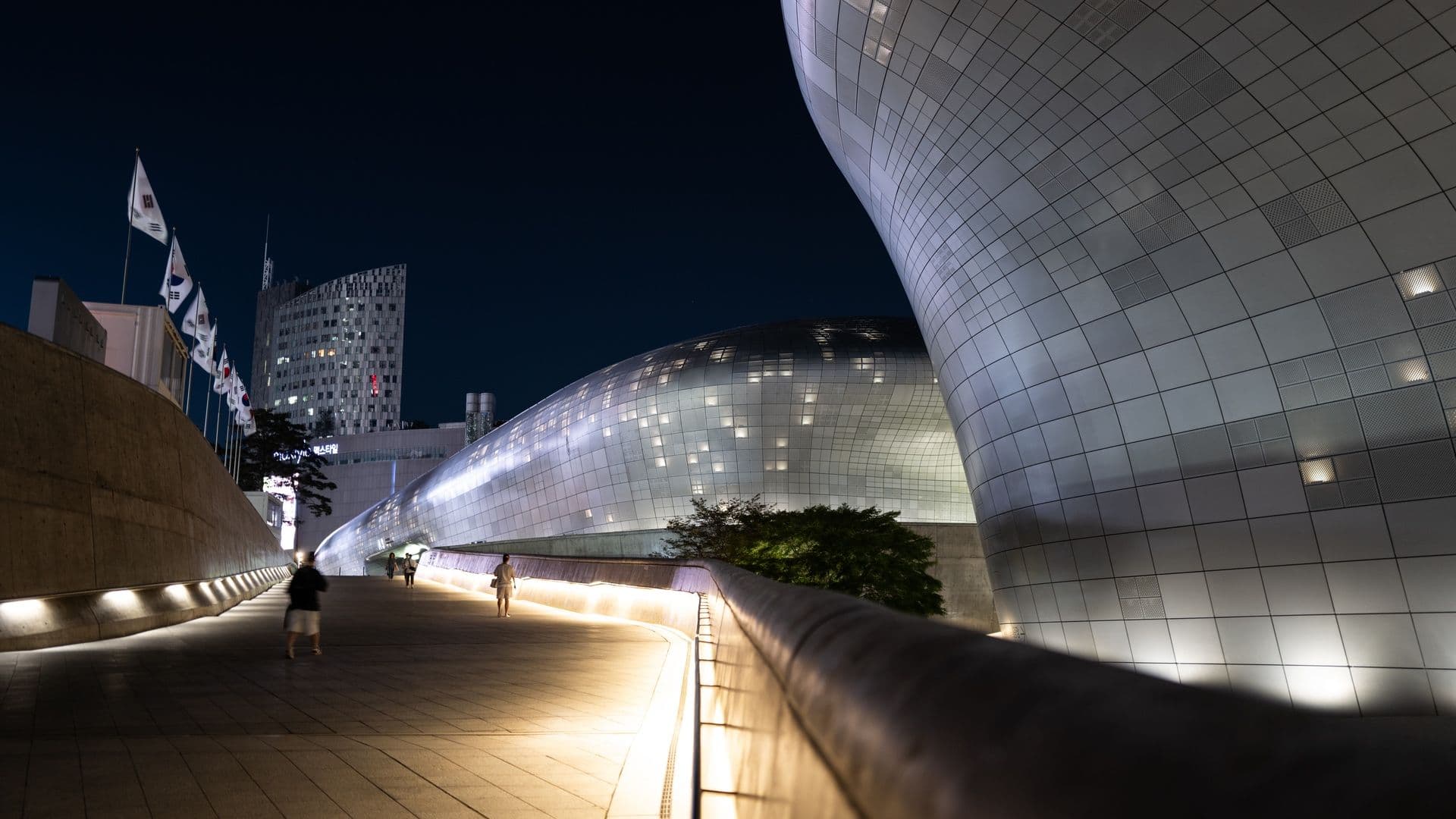 Night view of modern architectural buildings with curved metallic exteriors, illuminated walkways, and flags in South Korea.