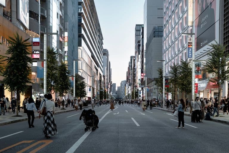 A busy city street with tall buildings on either side, many pedestrians walking, some wearing masks, and trees lining the sidewalks.