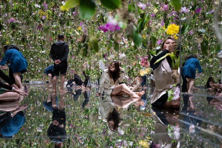 People in an indoor floral exhibit with hanging orchids, sitting and walking on a reflective wet floor, creating mirror reflections of the flowers and visitors.