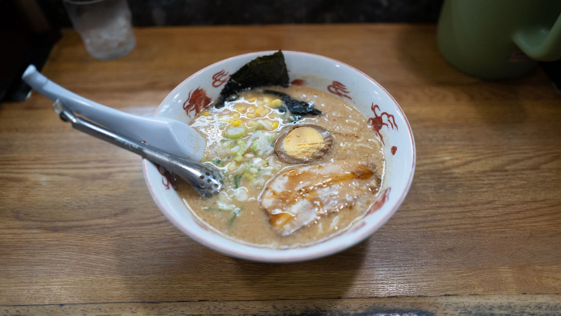 Bowl of ramen with broth, noodles, sliced pork, boiled egg, seaweed, corn, and green onions on a wooden table.